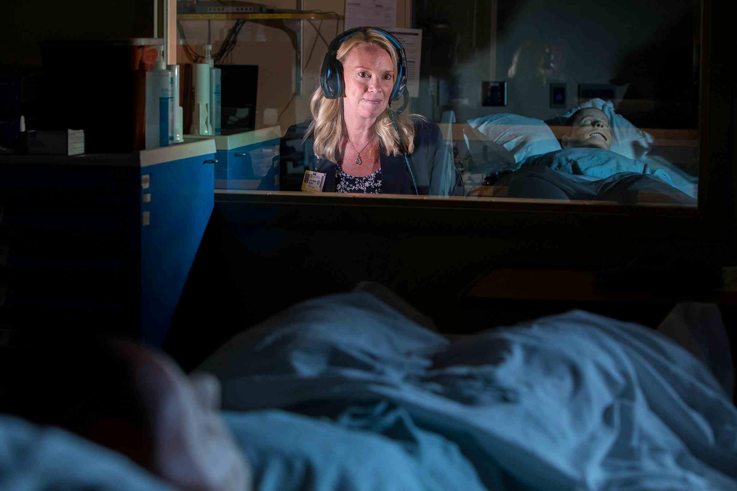 Erin Baylor wears a headset, sitting behind the glass wall of a simulation lab control room.