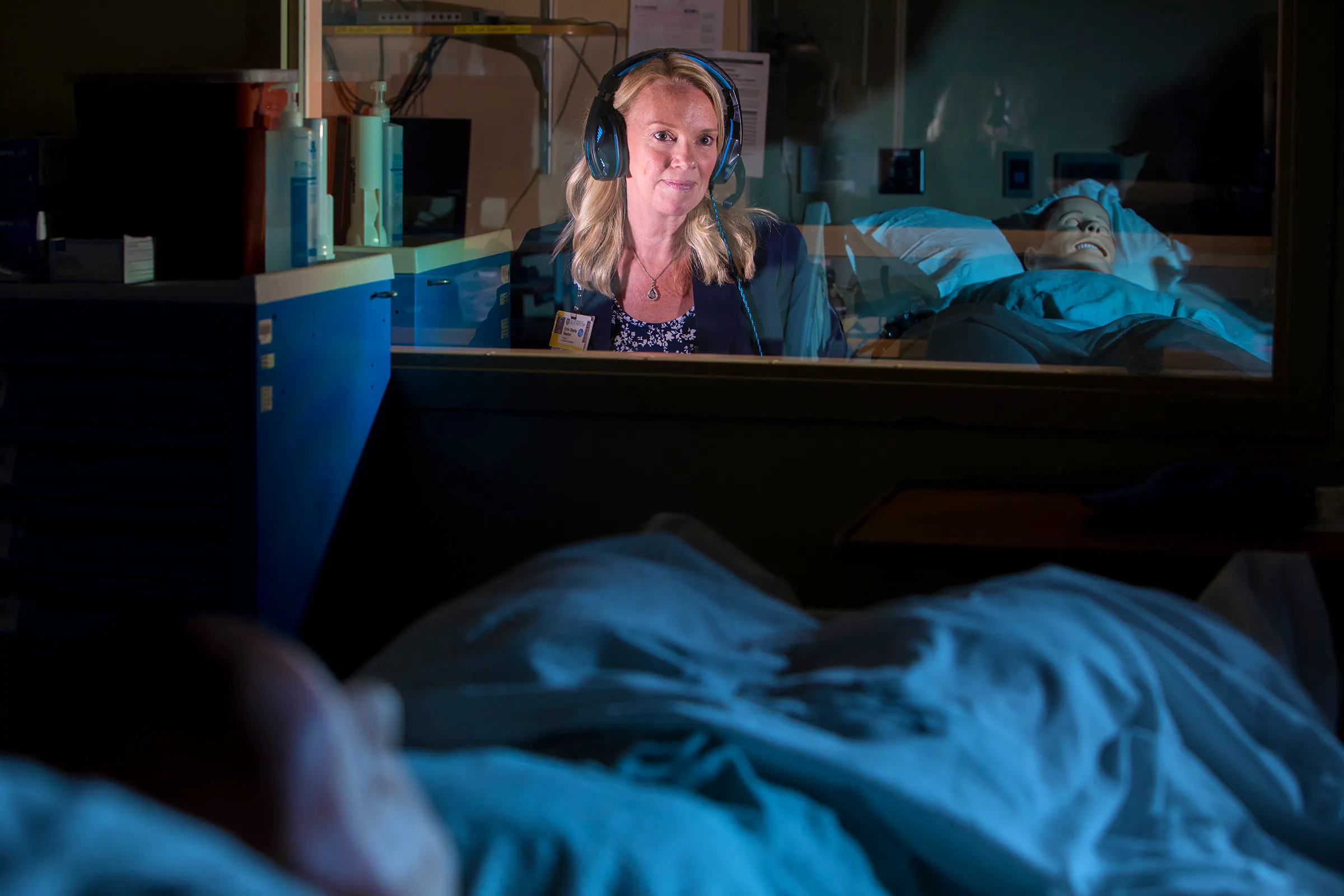 Erin Baylor wears a headset, sitting behind the glass wall of a simulation lab control room.