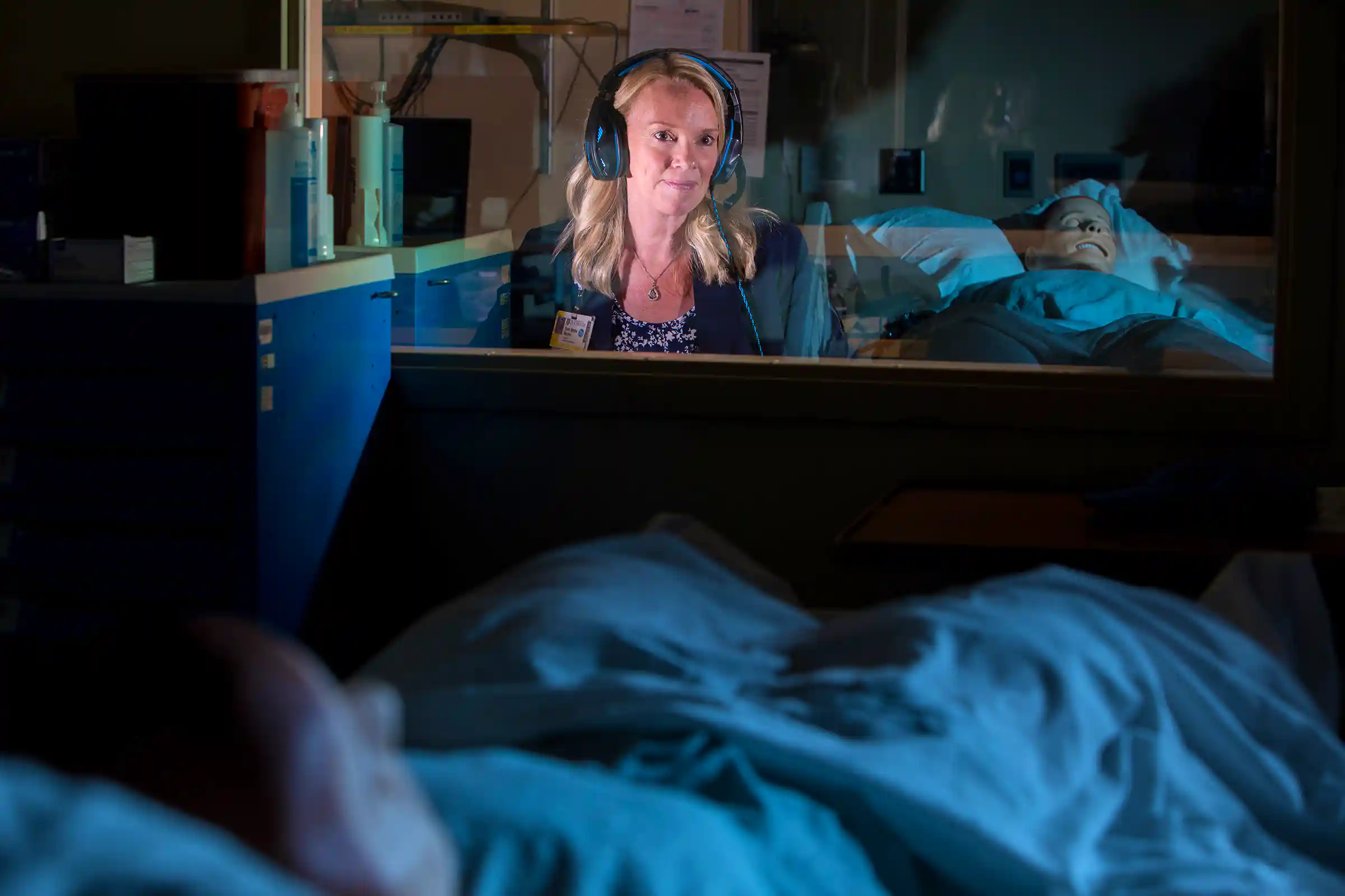 Erin Baylor wears a headset, sitting behind the glass wall of a simulation lab control room.