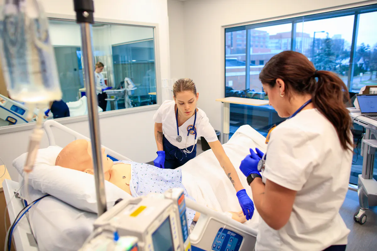 Two nursing students work together in a simulation suite, standing over a mannikin in a hospital gown.
