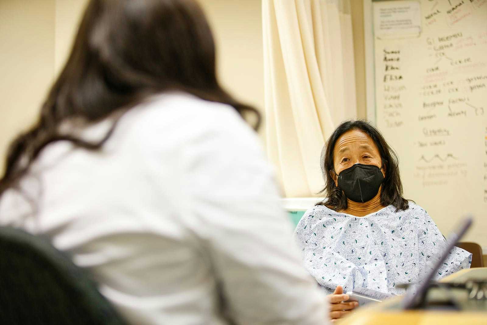Standardized patient with mask on looking at student.