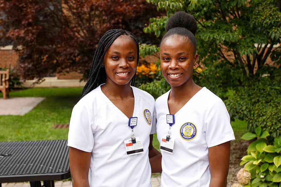 Twin sisters smiling in their nursing uniforms outside in the courtyard.