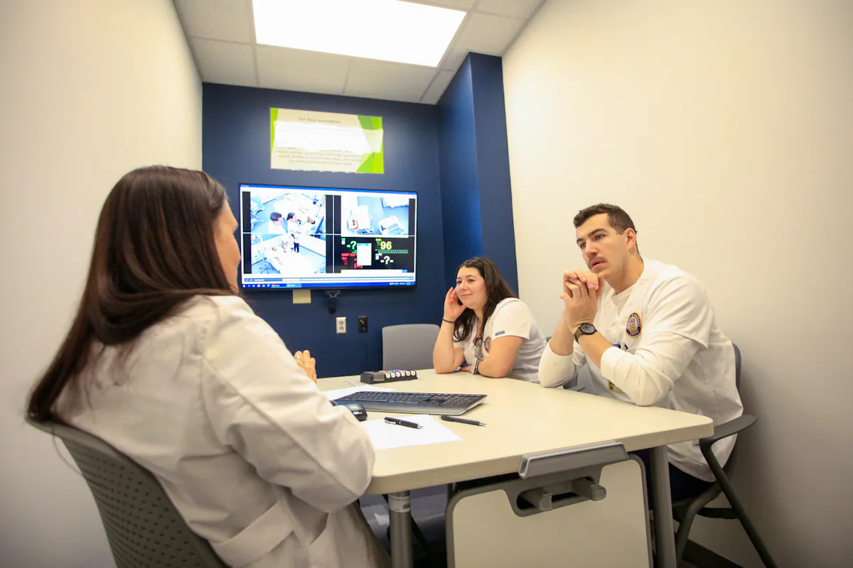 Students and professors speak at a debrief room table, with classroom video footage on a TV screen above them.