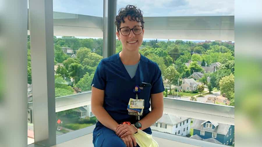 Erin Sabo sits in front of a window overlooking the University of Rochester Medical Center campus, wearing navy blue scrubs.
