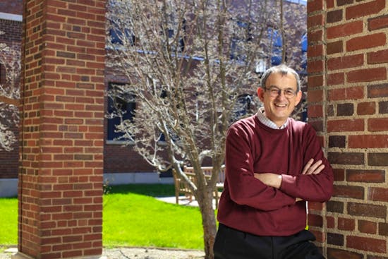 Patrick Hopkins stands next to a brick wall in the School of Nursing courtyard, wearing a read sweater and white plaid shirt underneath.