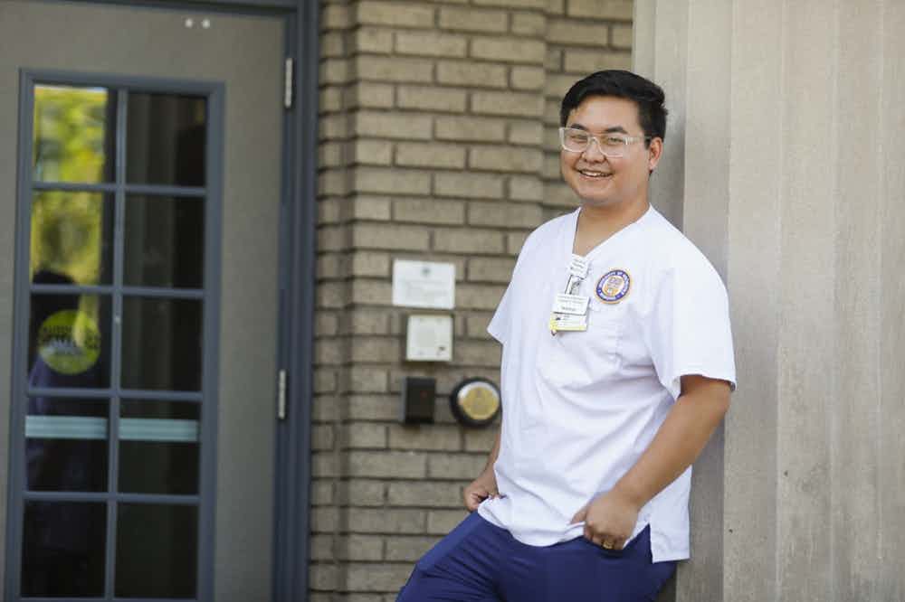 Thaw Htoo '23N stands outside the Children's School wearing his white School of Nursing scrubs.