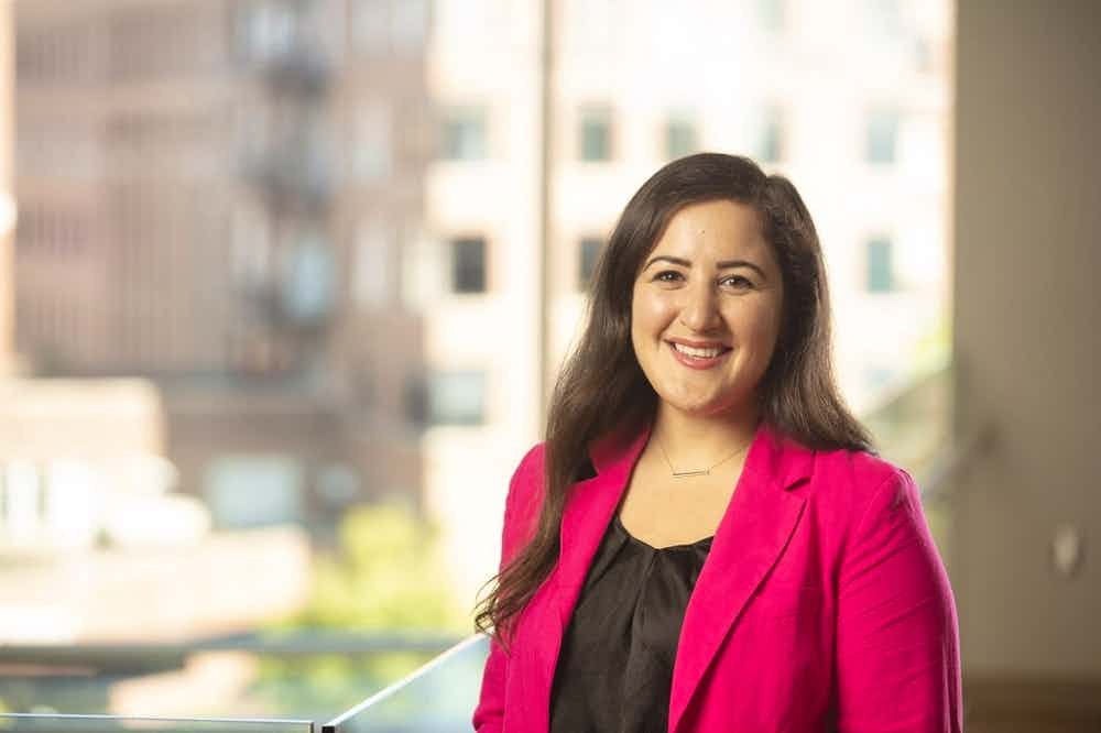 Portrait of Lauren Ghazal next to a window, wearing a black shirt and pink jacket.