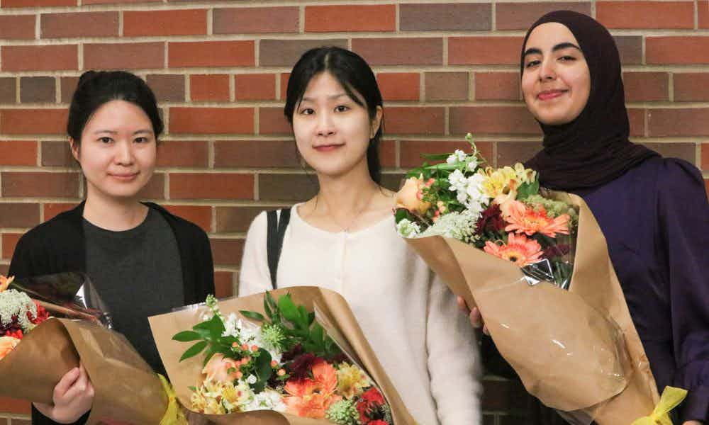 Three students hold bouquets of flowers in the School auditorium.