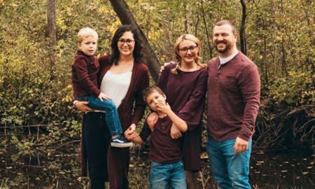 Family photo of Cassie Weis, husband Nicholas, and three children standing in front of green trees.
