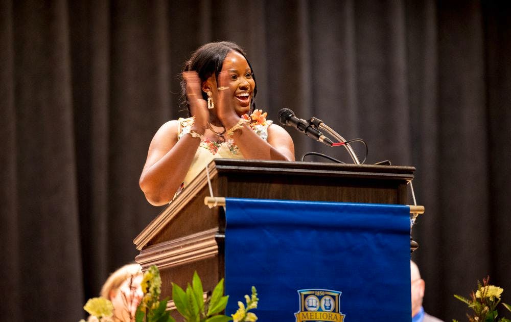 Student speaker smiles and applauds during a graduation speech while standing at a podium.