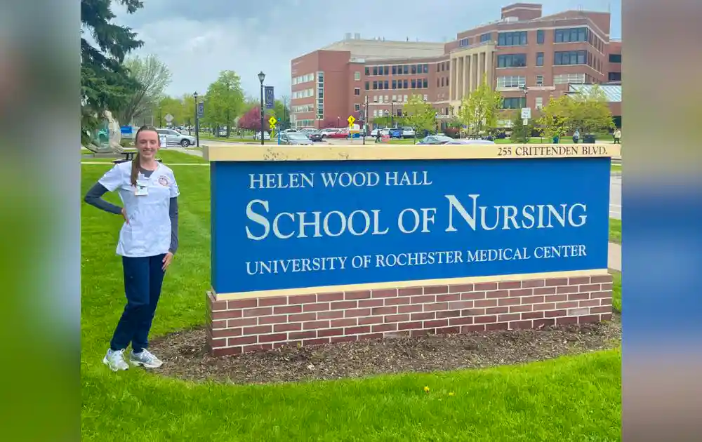 MaryGrace stands next to the School of Nursing sign on the front lawn. She is wearing the white and navy ABSN uniform.