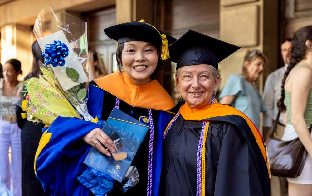 Two faculty members smile, holding flowers while standing outside Eastman Theatre.