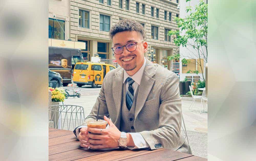 Man sitting at a table and holding an iced coffee, with city buildings behind him. 