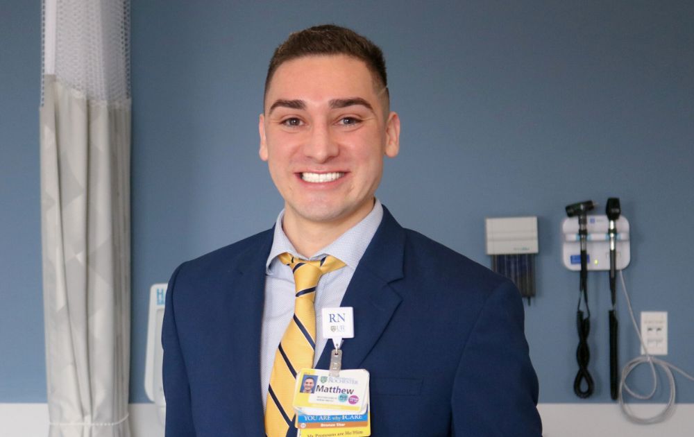 Photo of Matt standing in a patient room, wearing a navy jacket and yellow and blue striped tie.