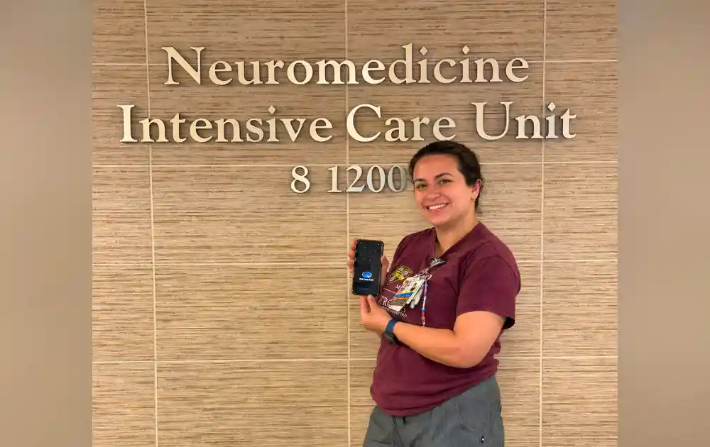 Caris stands in front of the sign for Strong's Neuro ICU holding up her phone with the Night Shift Mode App open.