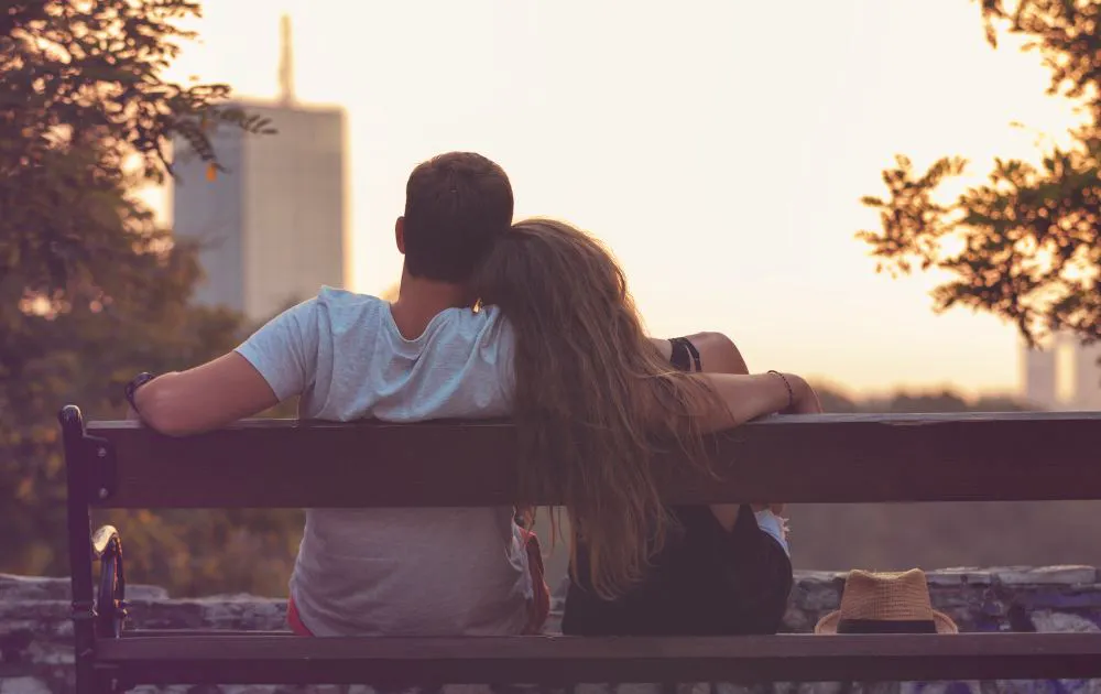 Photo of a man and woman sitting on a park bench, shown from behind.