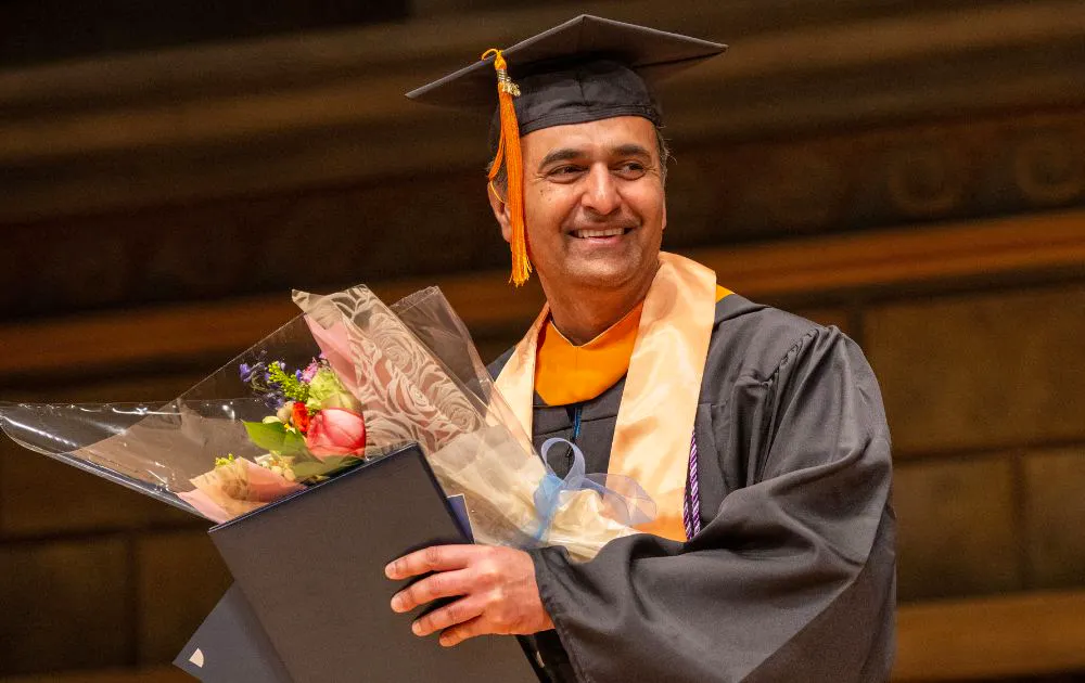 Graduate Bhavesh Shah accepts an award and flowers. He is smiling and wearing a black graduation cap and gown.