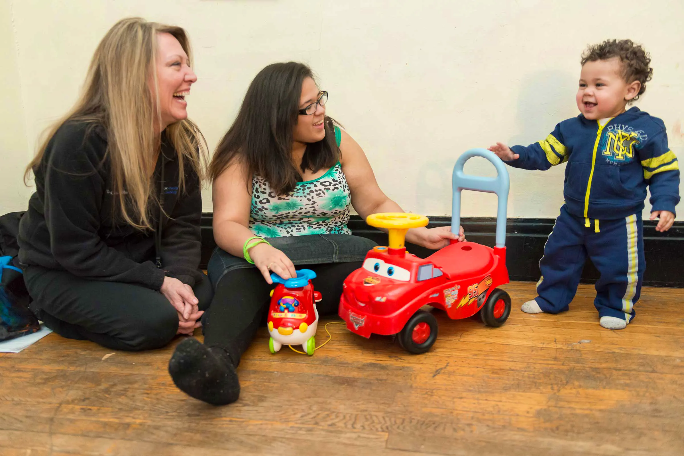 Two women smiling at a toddler who is walking towards his toy motorcycle