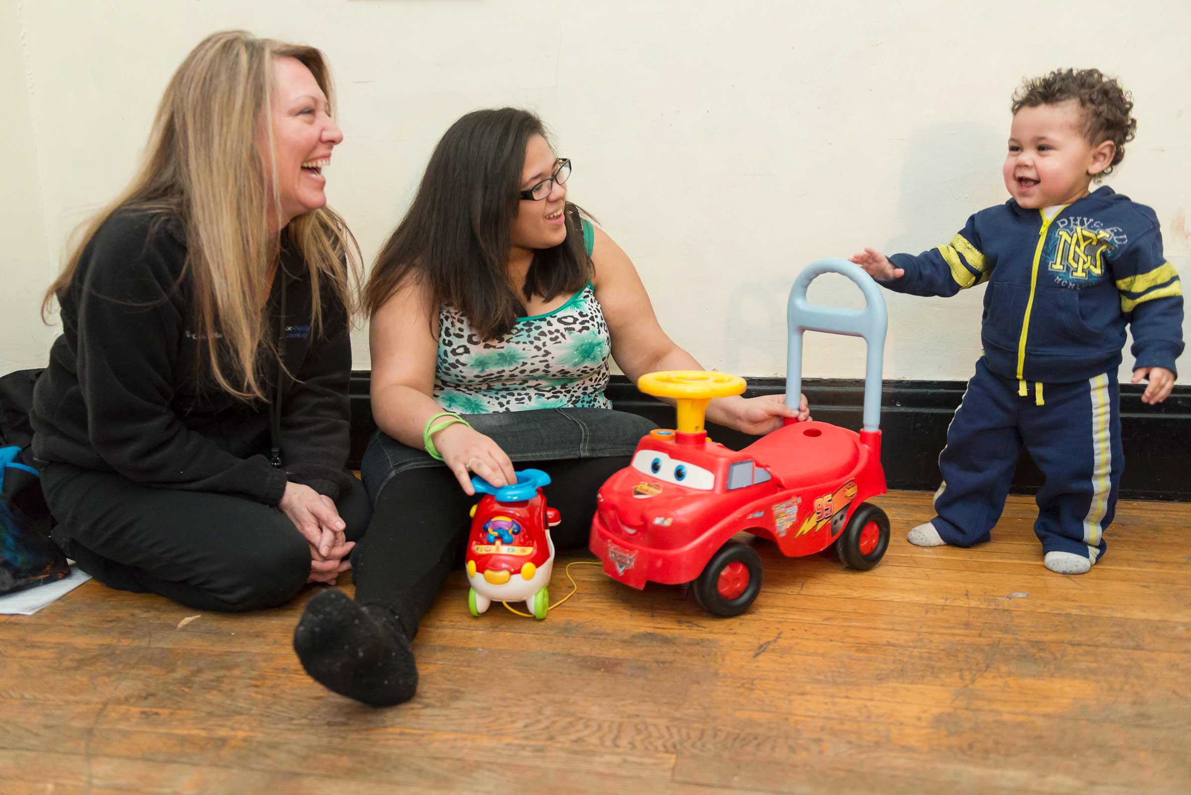 Two women smiling at a toddler who is walking towards his toy motorcycle